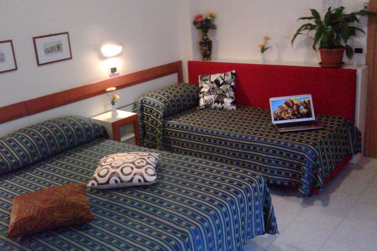 A hotel room with two beds, a red accent wall, a small table with a lamp, and a laptop on one bed.