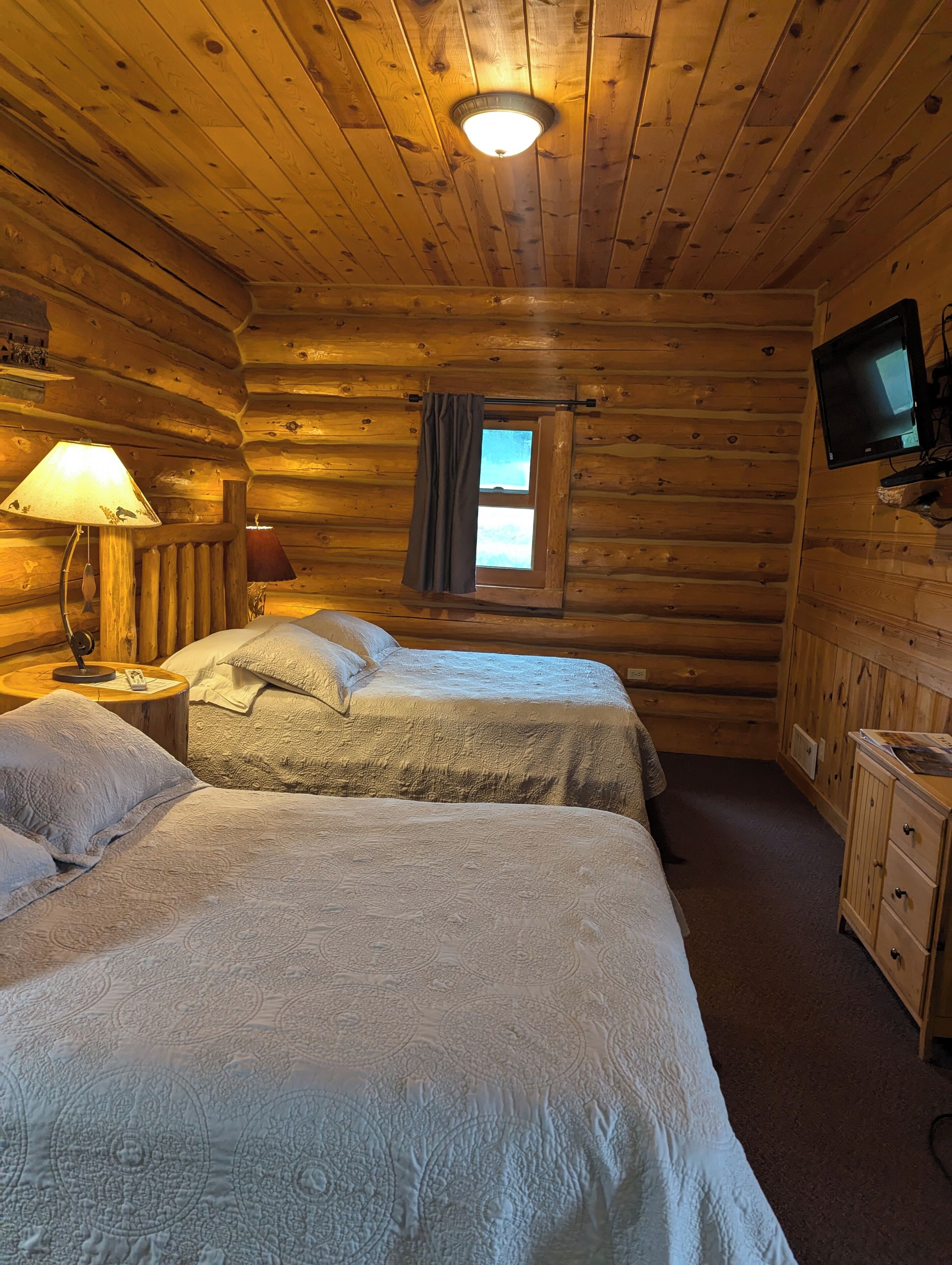Ein Schlafzimmer einer Blockhütte mit zwei Betten, einem Fernseher, einem Fenster mit Vorhängen und einer Lampe.