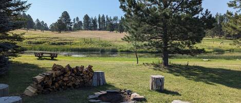 A serene outdoor setting with a picnic table, a stack of firewood, and a fire pit surrounded by trees and a grassy field.