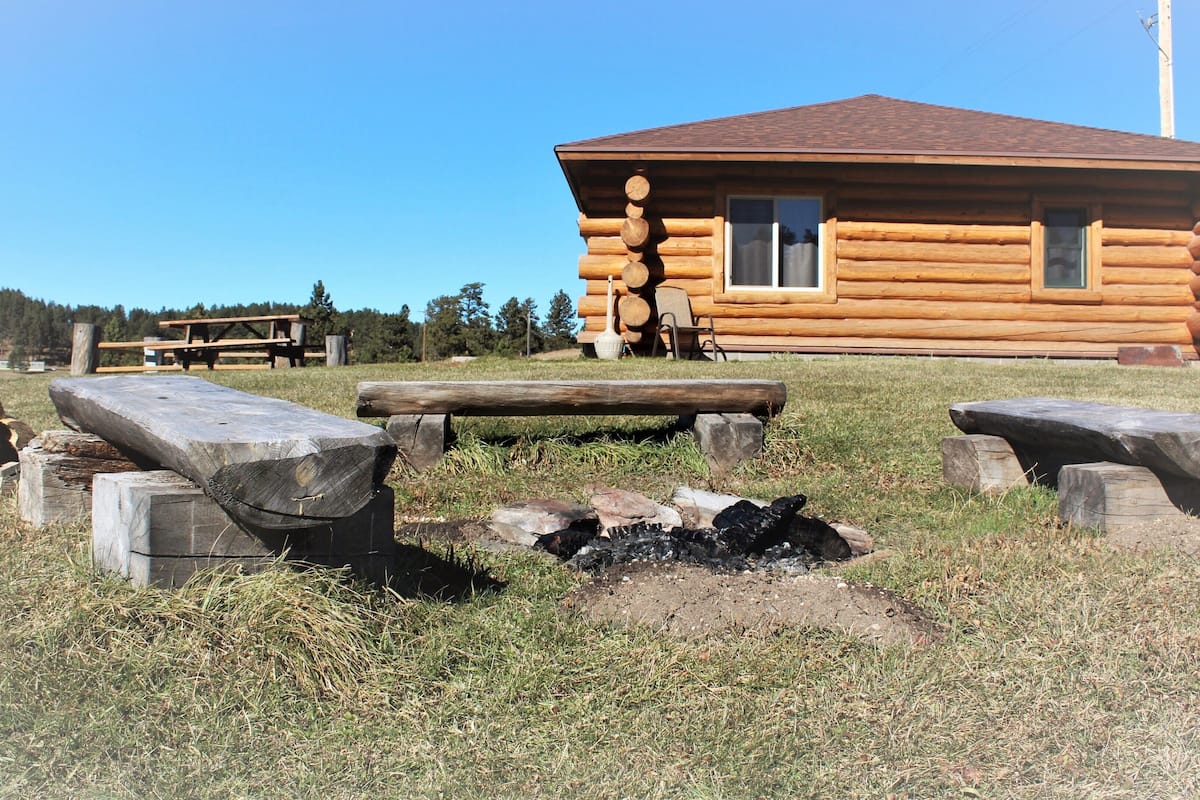 A log cabin with a fire pit and picnic table in a grassy area.