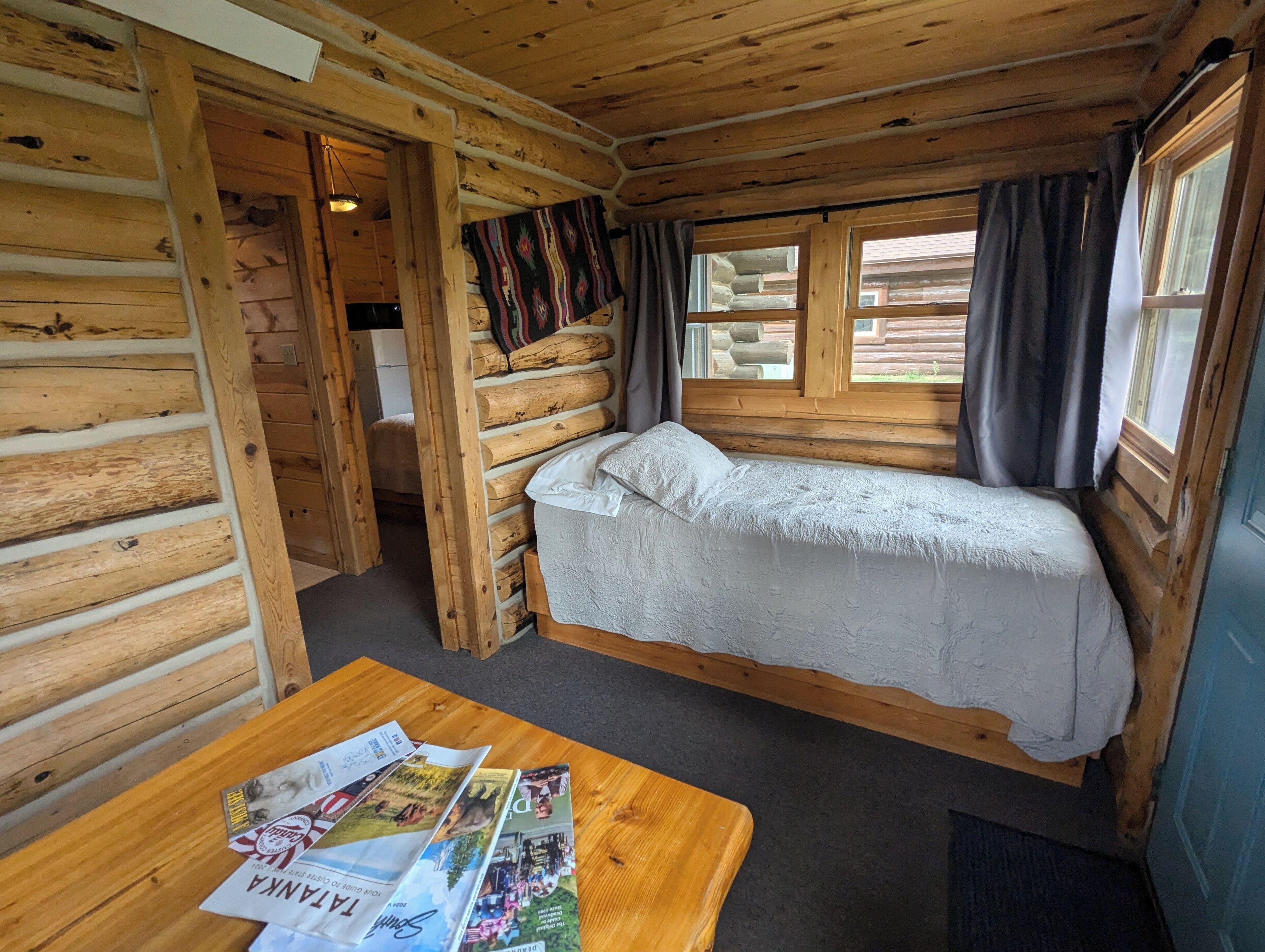 Een houten blokhutkamer met een bed, een tafel met tijdschriften en een raam met gordijnen.