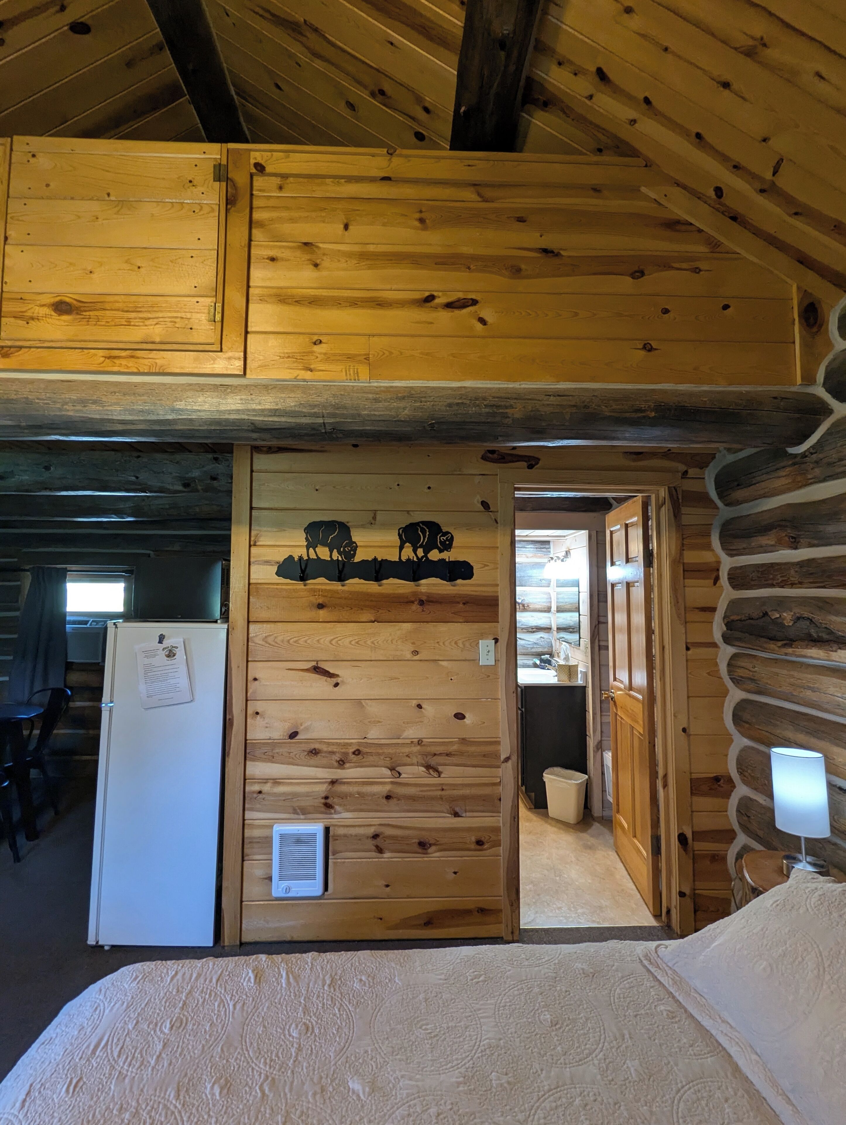 A log cabin interior with a bed, a refrigerator, a sink, and a wooden ceiling.
