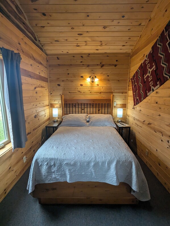 Une chambre dans un chalet en bois, avec un lit, deux tables de chevet, un luminaire fixé au mur et une fenêtre avec des rideaux.
