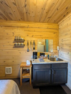 Une cuisine compacte aux murs en bois, dotée d’un plan de travail en granit et d’un évier. On y trouve des étagères avec des tasses et une machine à café.