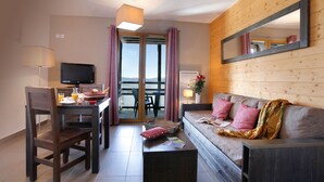 A hotel room with a wooden interior, a sofa with cushions, a dining table with chairs, a TV, and a view through a glass door. - Apart-Hôtel Le Pic de l'Ours (Font-Romeu-Odeillo-Via)