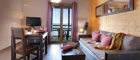 A hotel room with a wooden interior, a sofa with cushions, a dining table with chairs, a TV, and a view through a glass door.