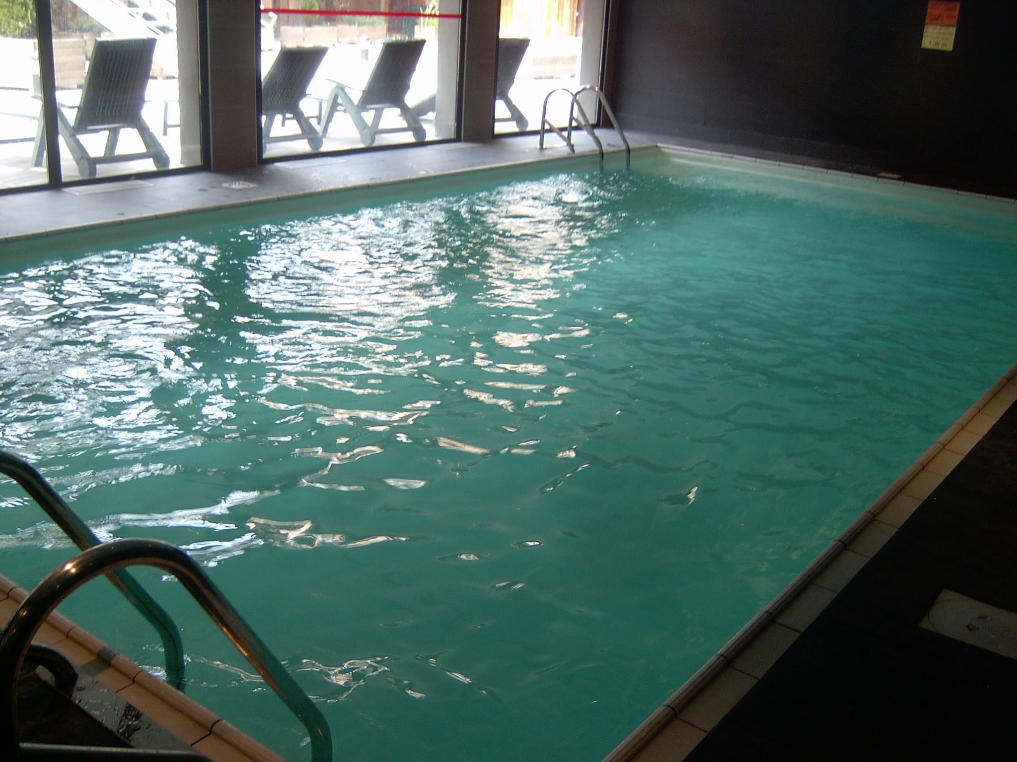 Une piscine intérieure à l’eau claire, avec un escalier et des chaises longues disposées à l’extérieur, devant la fenêtre.