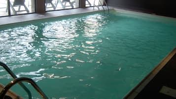Une piscine intérieure à l’eau claire, avec un escalier et des chaises longues disposées à l’extérieur, devant la fenêtre.