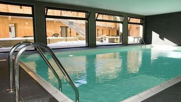 An indoor swimming pool with clear glass walls, metal ladders, and a tiled floor. - Apart-Hôtel Le Pic de l'Ours (Font-Romeu-Odeillo-Via)