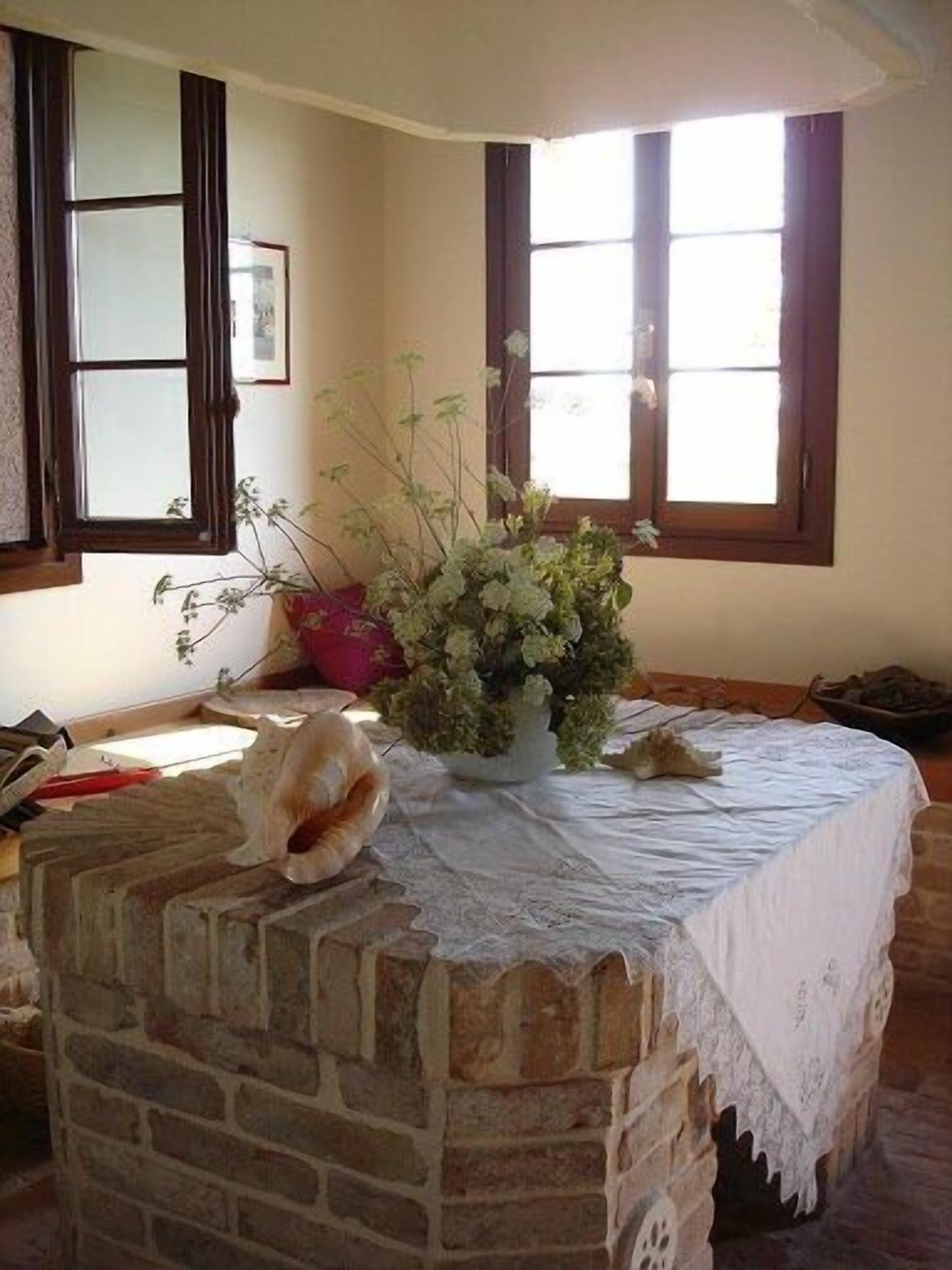 Une pièce rustique avec une cheminée en pierre, une table recouverte d’une nappe en dentelle et une fenêtre avec des volets en bois.