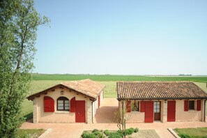Dos edificios rústicos con contraventanas rojas, rodeados de campos verdes bajo un cielo despejado.