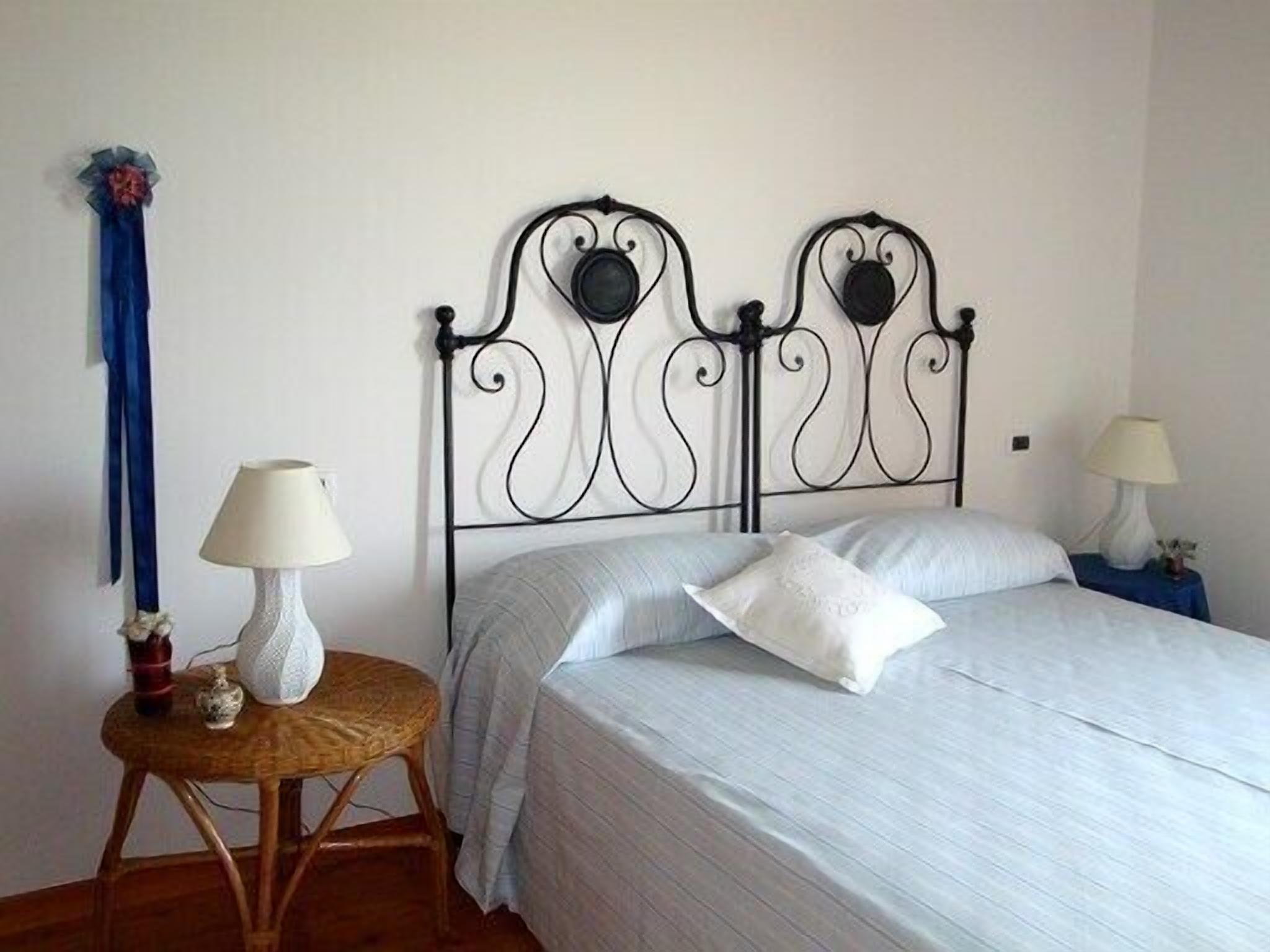 Quarto bem arrumado com uma cama, duas mesas de cabeceira com abajures, uma cadeira e um laço azul decorativo na parede.
