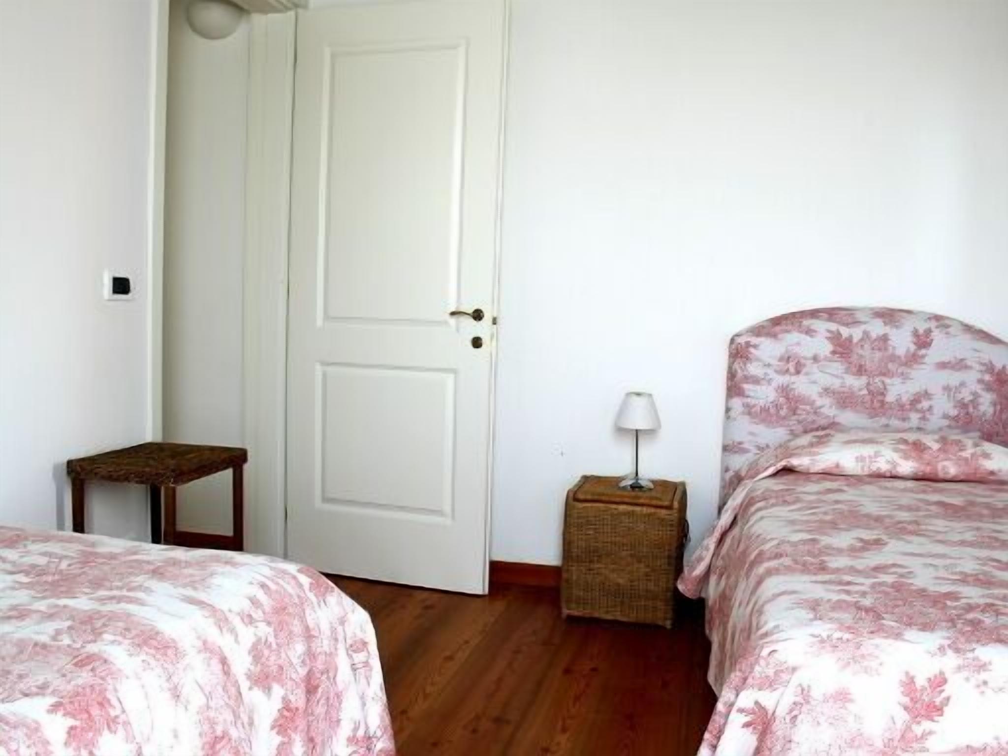 A bedroom with two beds, a white door, a small table, a lamp, and a wicker basket.