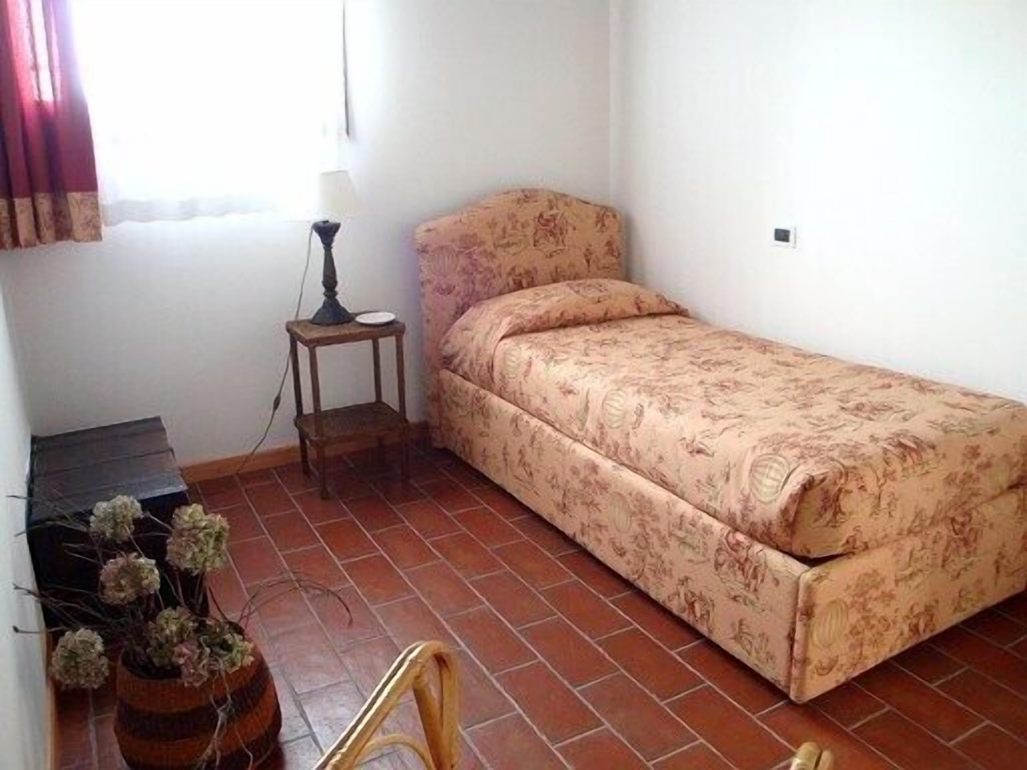 A small, single-bed room with a patterned bedspread, a small wooden nightstand with a lamp, and a potted plant.