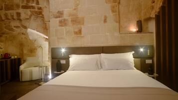 A hotel room with a large bed, a headboard with lights, a stone wall, a small table with a vase, and a chair.