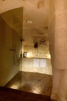Una stanza stretta e scarsamente illuminata, con zona doccia, un lavandino piccolo e una parete dalla superficie ruvida.