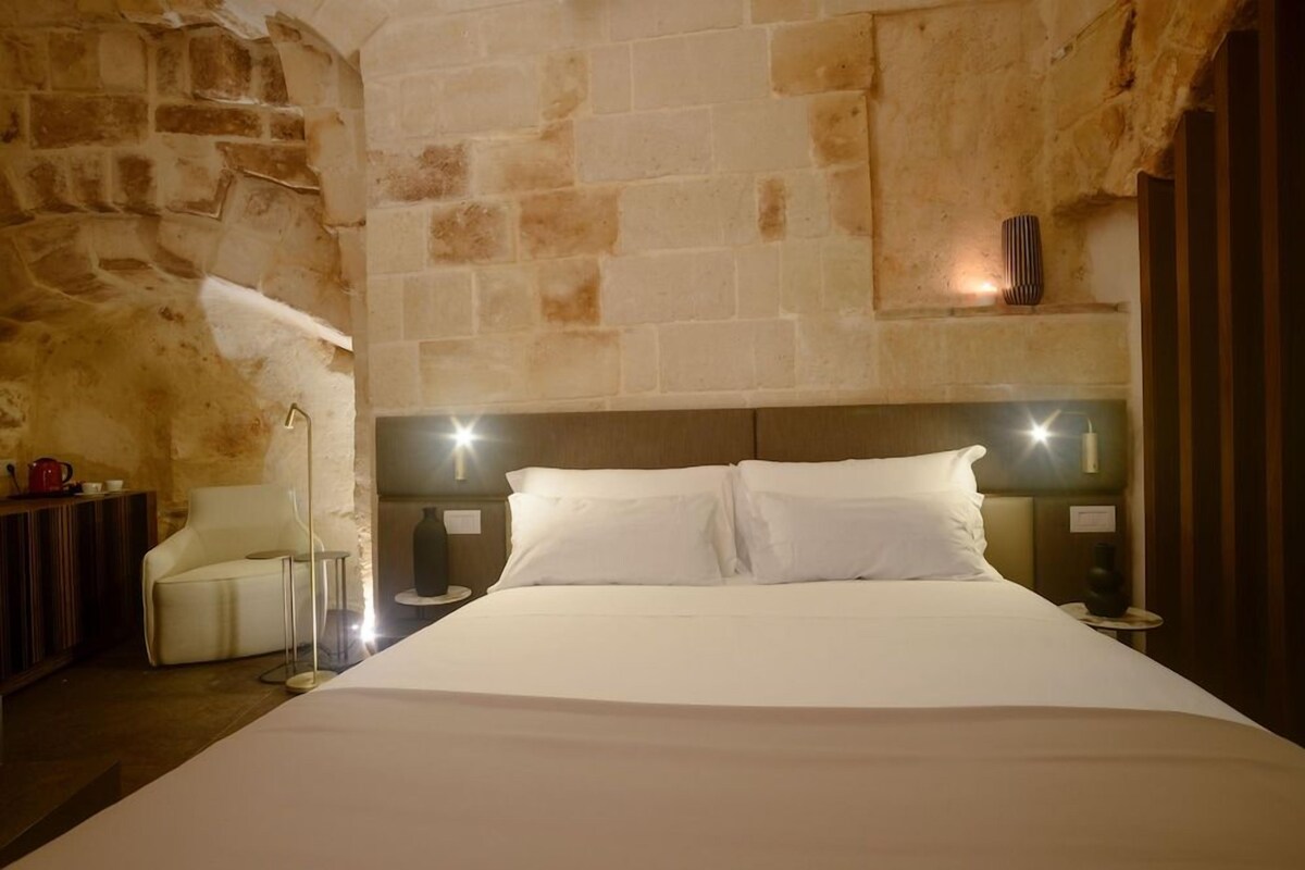 Une chambre d’hôtel avec un mur en pierre, un lit avec du linge de lit blanc, une table de chevet et une lampe.