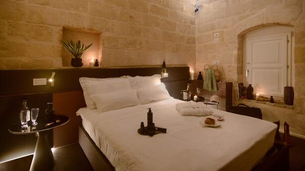 A hotel room with a stone wall, a bed with white linens, a bedside table with a bottle and glasses, a small plant, and a nightstand with a lamp. - Le Malve Cave Retreat (Matera)