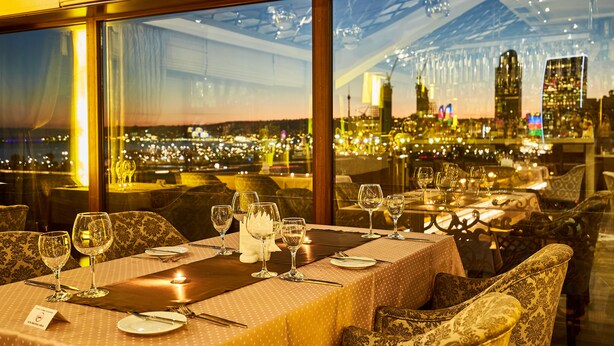 Een restaurant met gedekte tafels, met een stadsgezicht te zien door grote ramen.