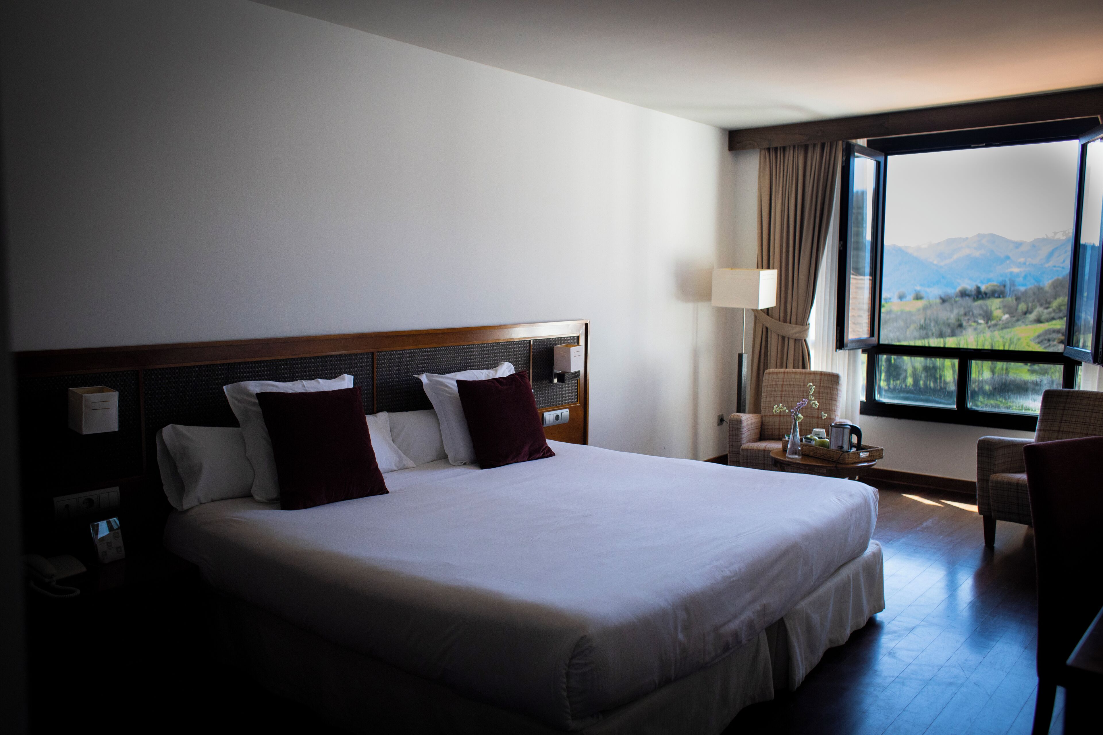 Habitación de hotel con una cama grande, una mesita de noche, una silla y vistas a las montañas por la ventana.
