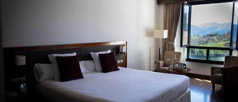 A hotel room with a large bed, a nightstand, a chair, and a view of mountains through the window.
