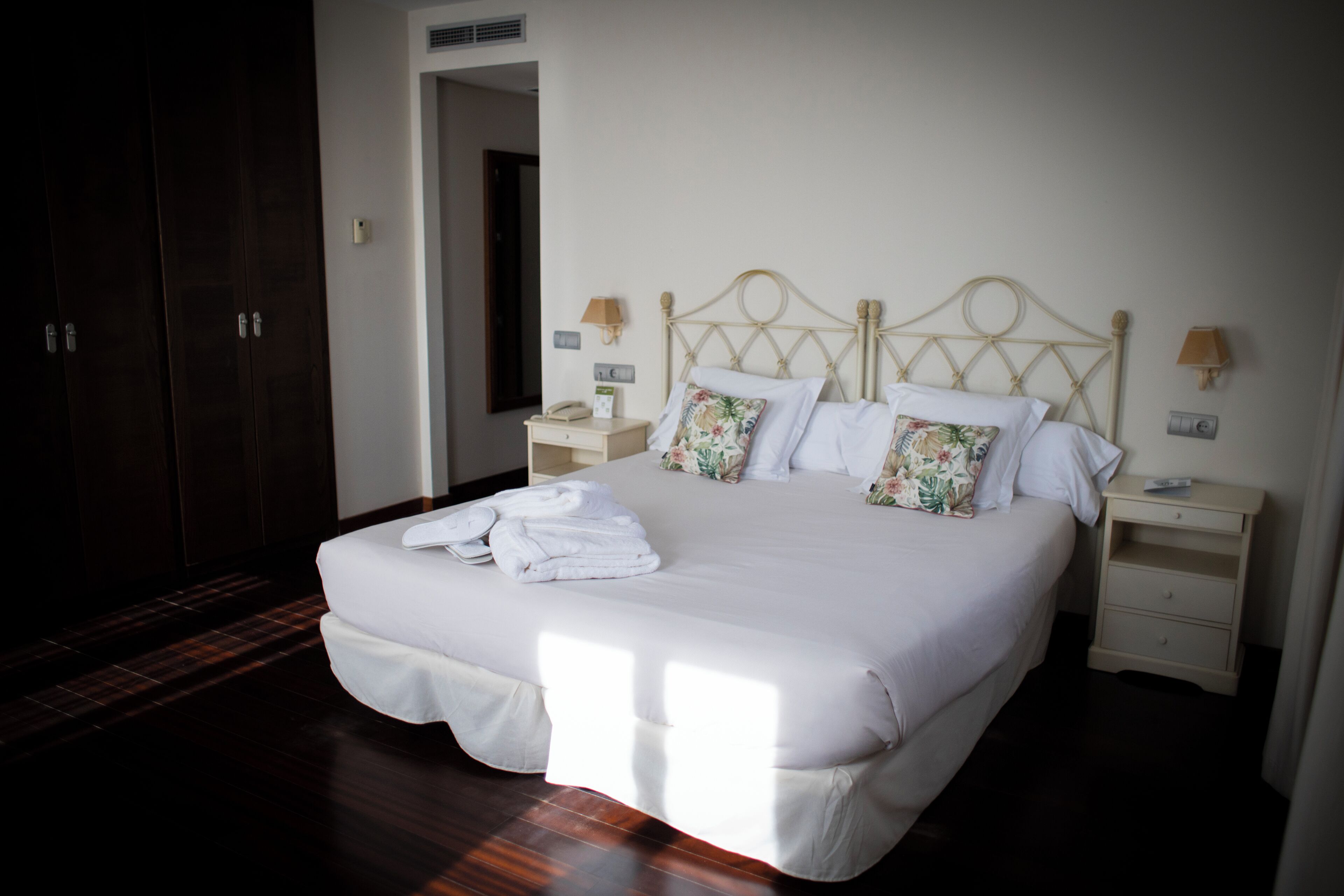 Una cama bien hecha con sábanas blancas y almohadillas florales, una mesita de noche con lámpara y un armario de madera oscura en una habitación de hotel.