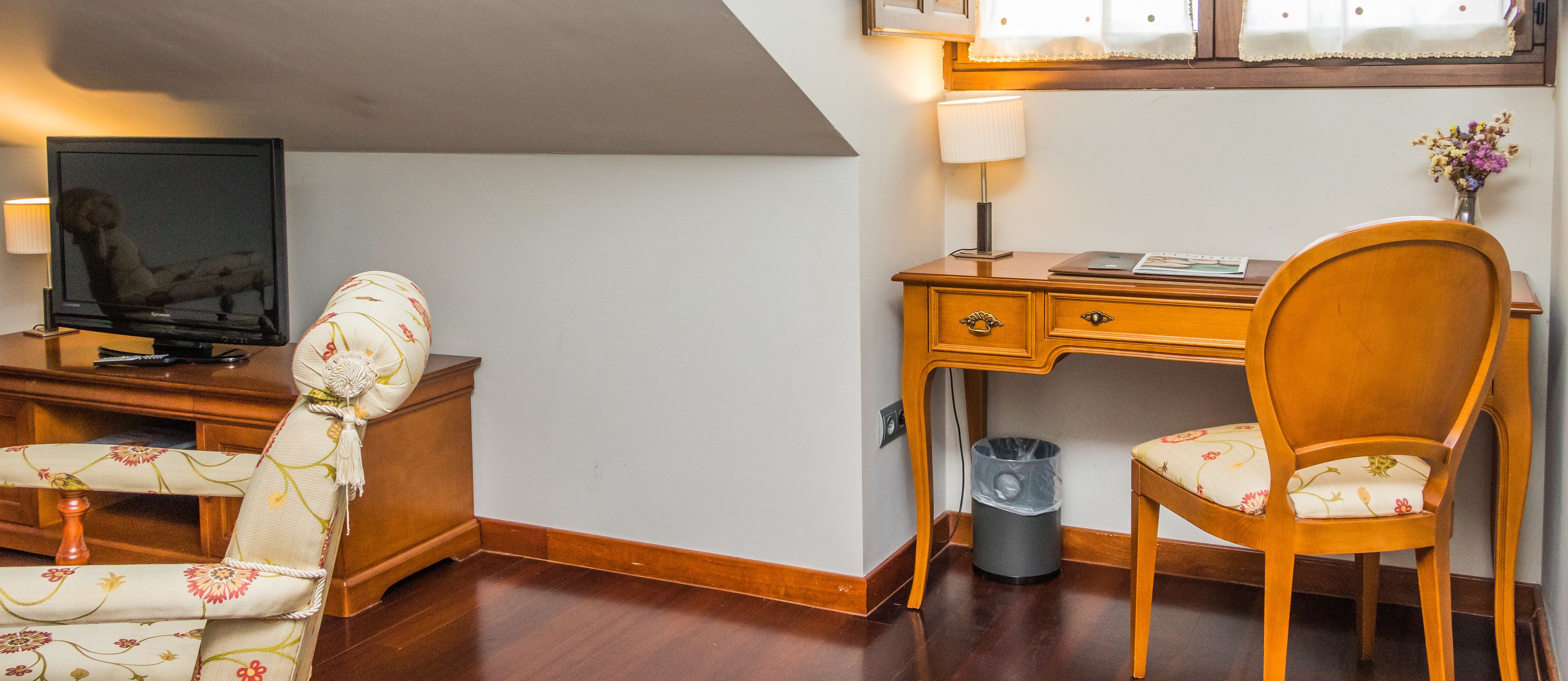 Una camera d'albergo con una scrivania in legno, una sedia, una televisione, una lampada e un cestino.