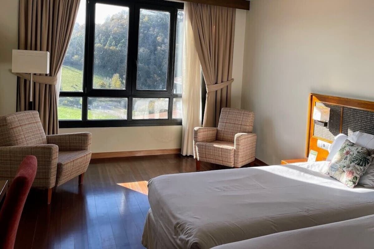 Une chambre d’hôtel avec un grand lit, deux fauteuils, une table de chevet et une fenêtre avec des rideaux.