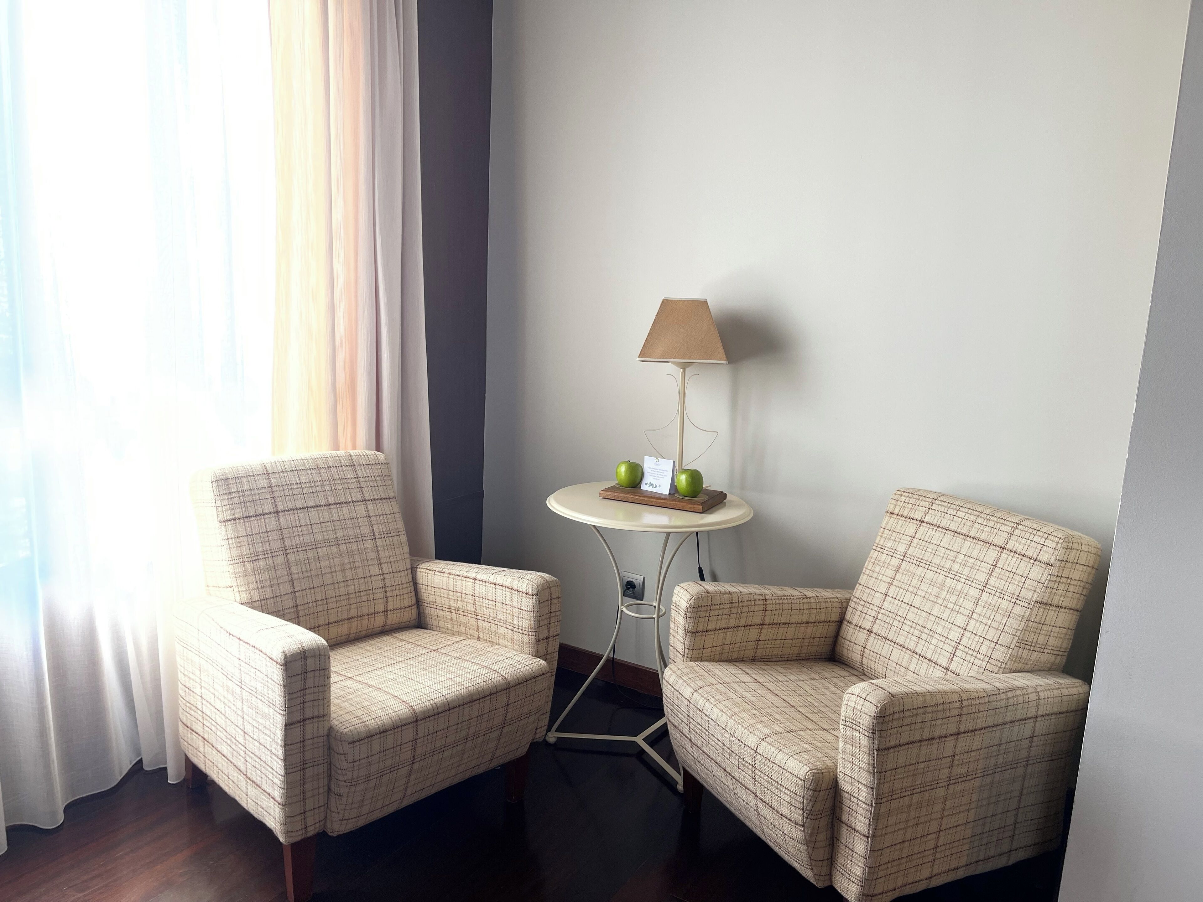 A hotel room with two plaid armchairs, a small round table with a lamp, and a view of the outdoors through sheer curtains.