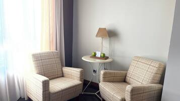 Habitación de hotel con dos sillones a cuadros, una mesita redonda con lámpara y vistas al exterior a través de cortinas translúcidas.