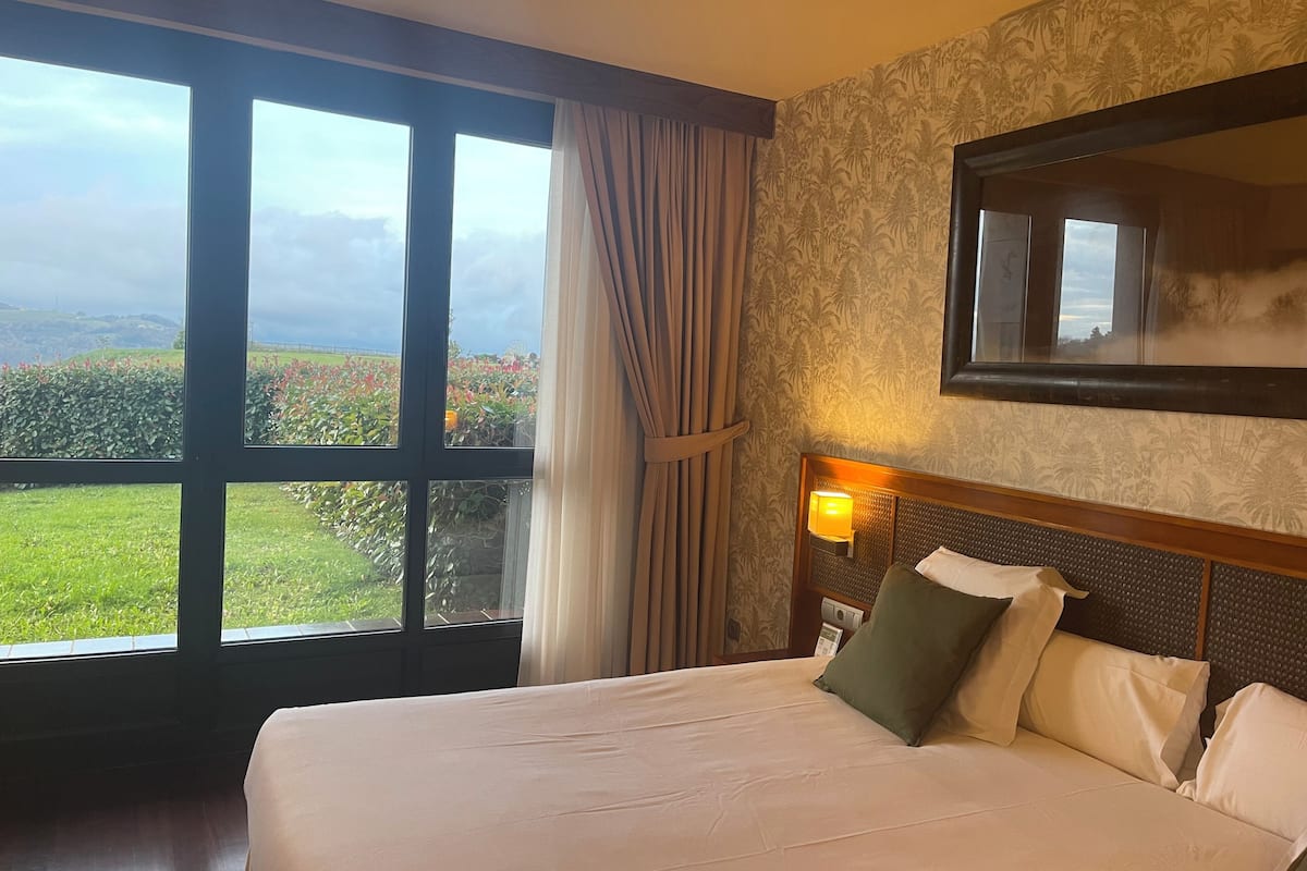 Une chambre d’hôtel dotée d’un grand lit, d’une table de chevet, d’une lampe fixée au mur et d’une grande fenêtre donnant sur un paysage verdoyant et vallonné.