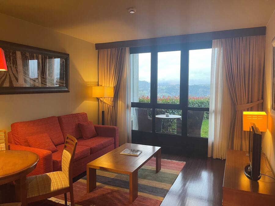 A hotel room with a red sofa, a wooden coffee table, a small round table with a cup, and a view of a garden through large windows.