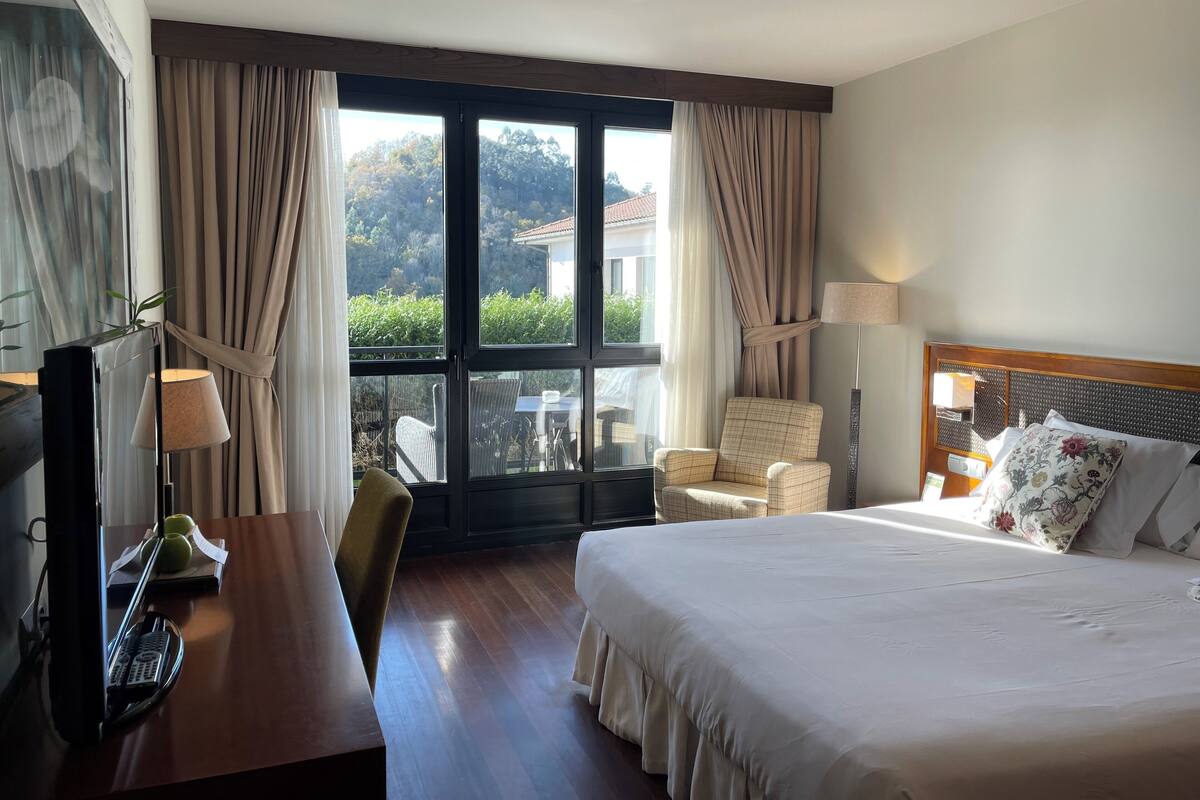 Une chambre d’hôtel avec un grand lit, un bureau équipé d’un ordinateur, une chaise et une vue sur la verdure environnante.