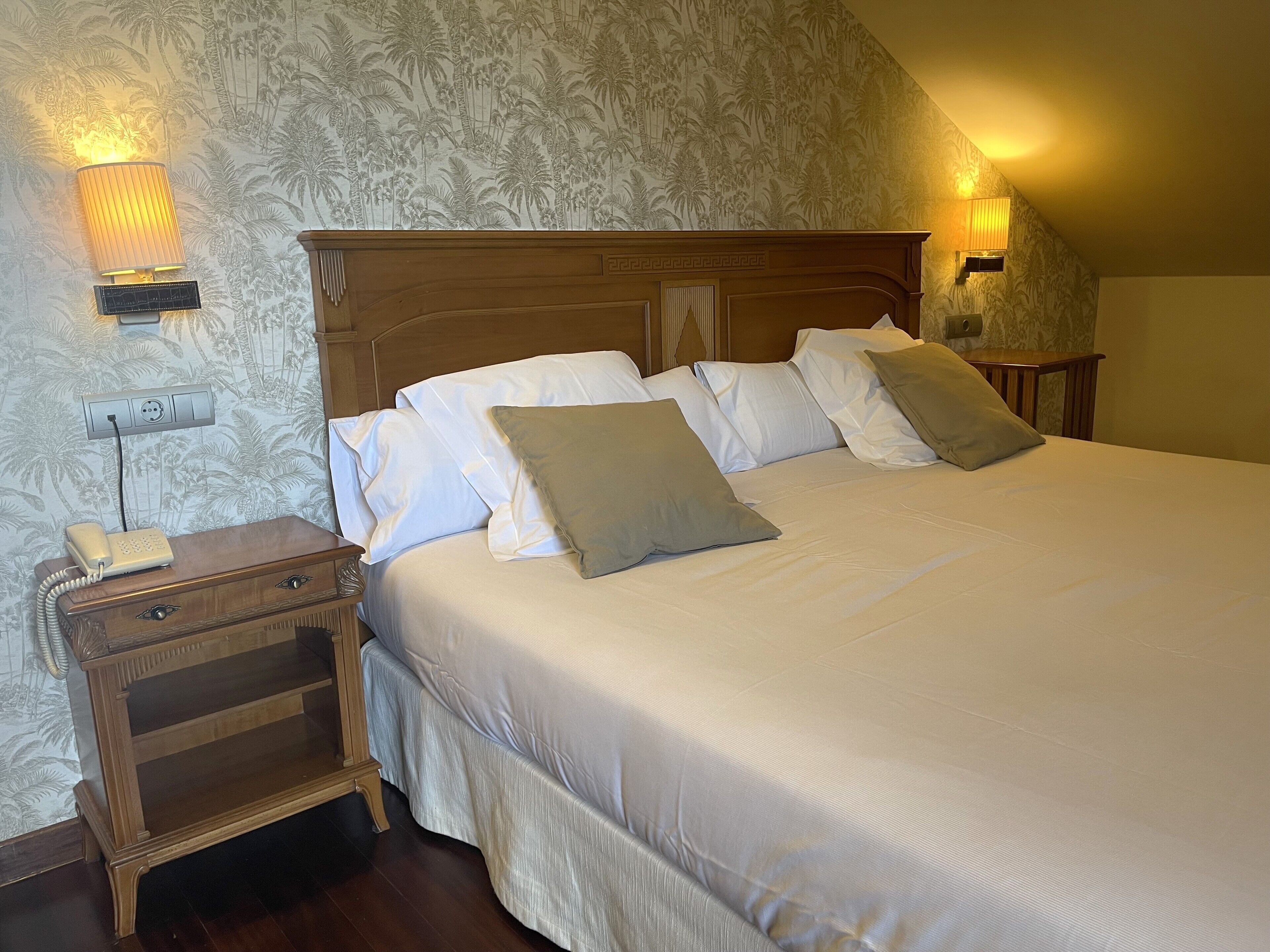 Ein ordentlich bezogenes Bett mit weißen Laken und zwei Kissen, ein Nachttisch aus Holz mit einem Telefon und eine Wandlampe.
