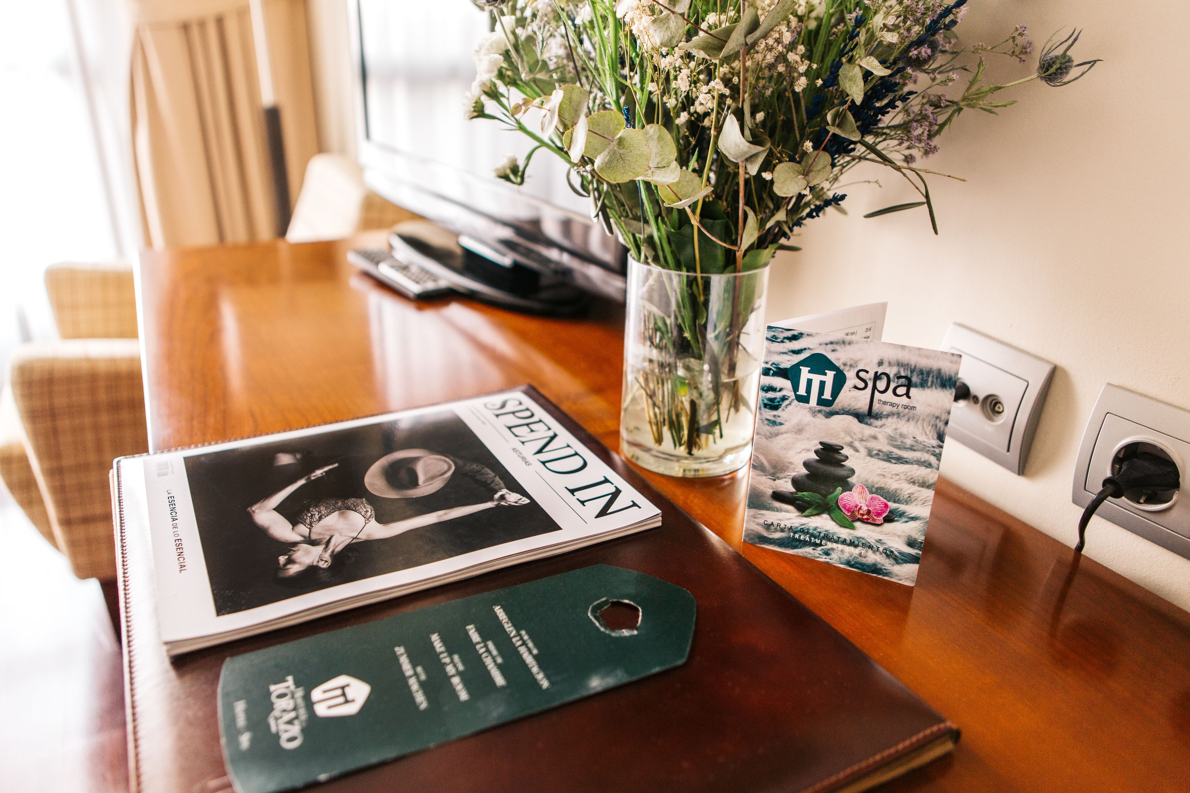 Et træbord med et magasin, en glasvase med blomster, en brochure for wellness og en hængeskilt.