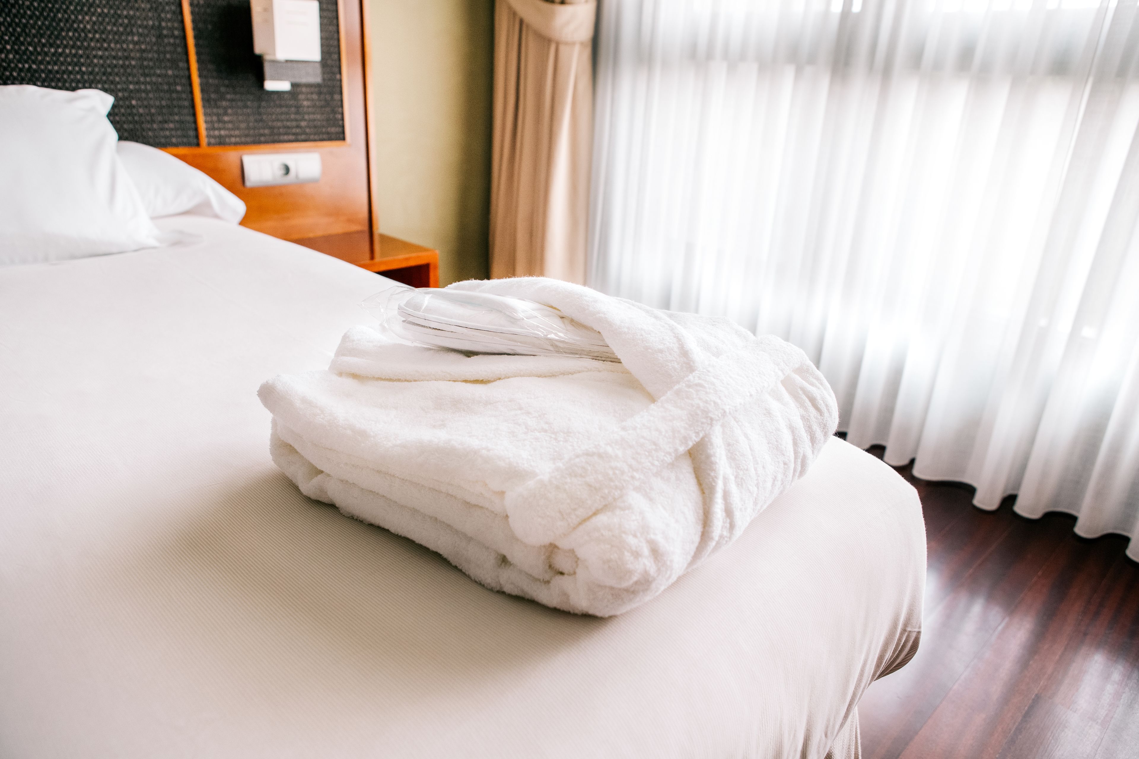 A neatly made bed with a folded white towel on top, a wooden nightstand with a lamp, and a window with sheer curtains.
