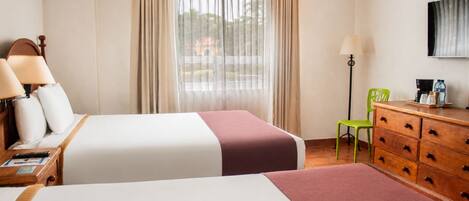 A hotel room with two beds, a desk with a lamp, a dresser, a chair, and a TV on the wall.