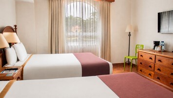 A hotel room with two beds, a desk with a lamp, a dresser, a chair, and a TV on the wall.