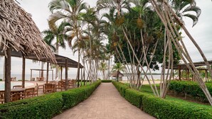 Un chemin pavé, bordé de haies soigneusement taillées et de palmiers, mène à une zone de plage aménagée avec des tables et des chaises en bois.