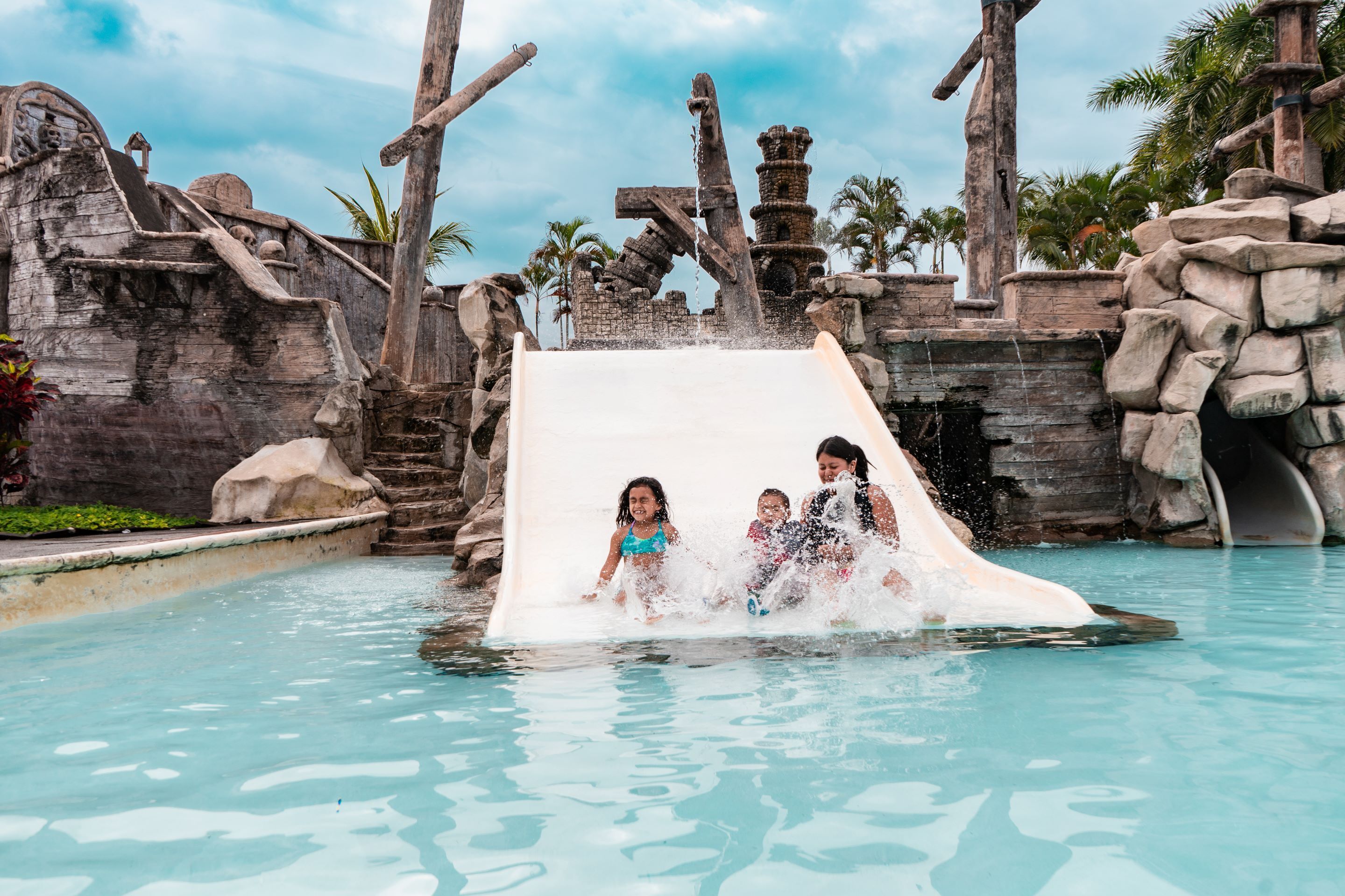 A water slide in a pool area with stone structures and palm trees.