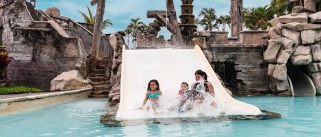 A water slide in a pool area with stone structures and palm trees.