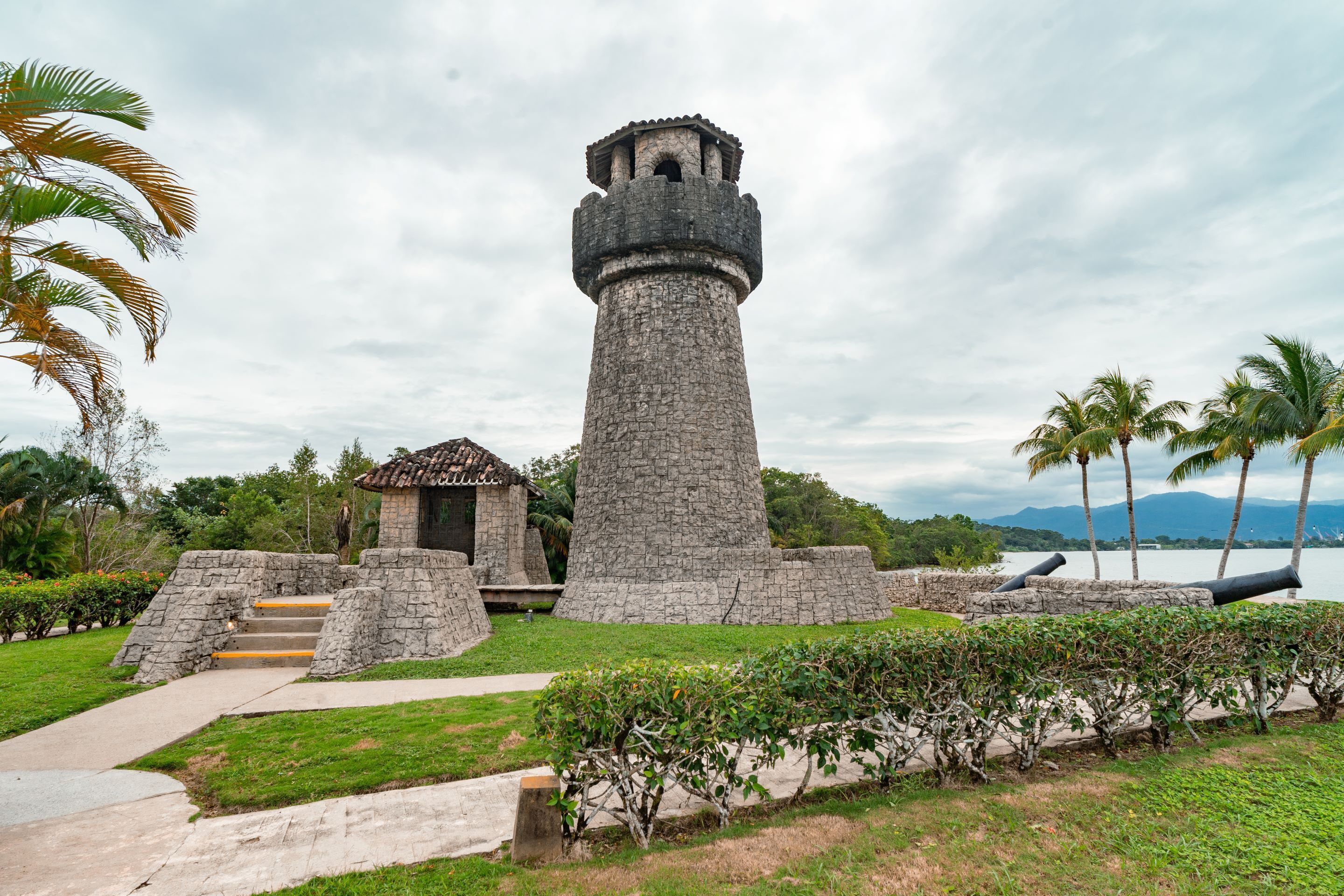 A stone lighthouse with a small adjacent structure, surrounded by a well-maintained garden and palm trees.