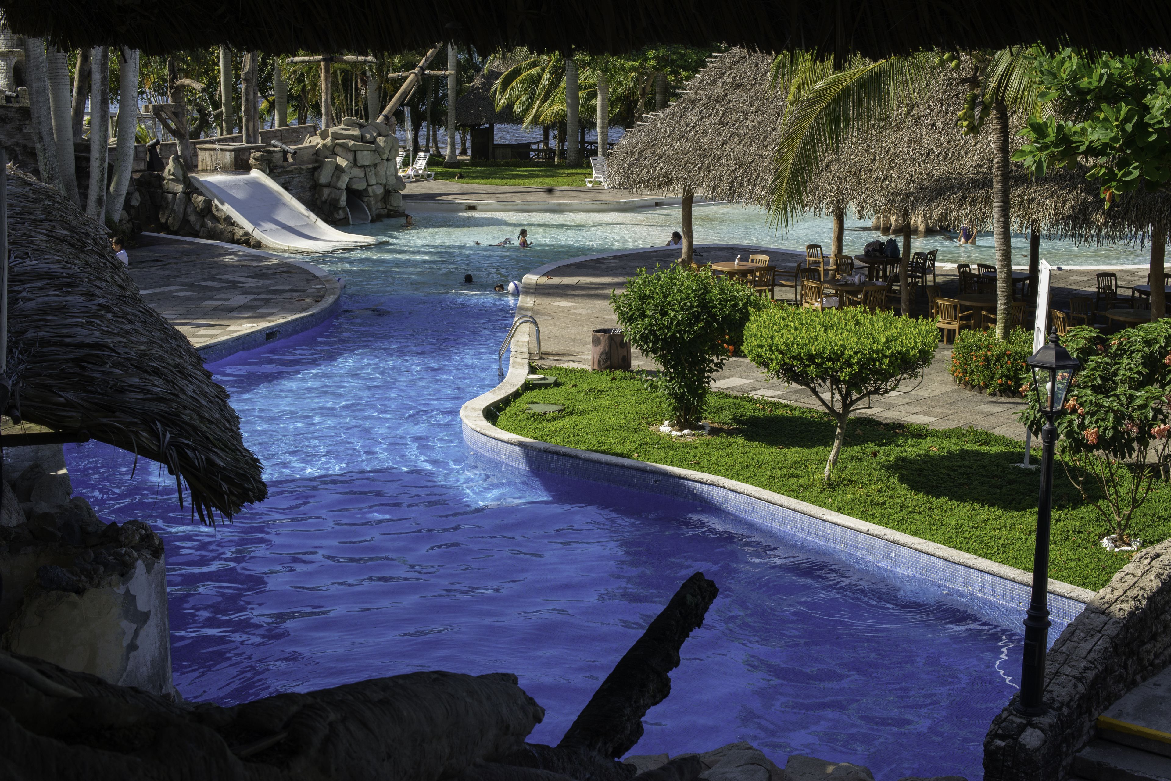A swimming pool with a slide, surrounded by palm trees and lounge chairs.