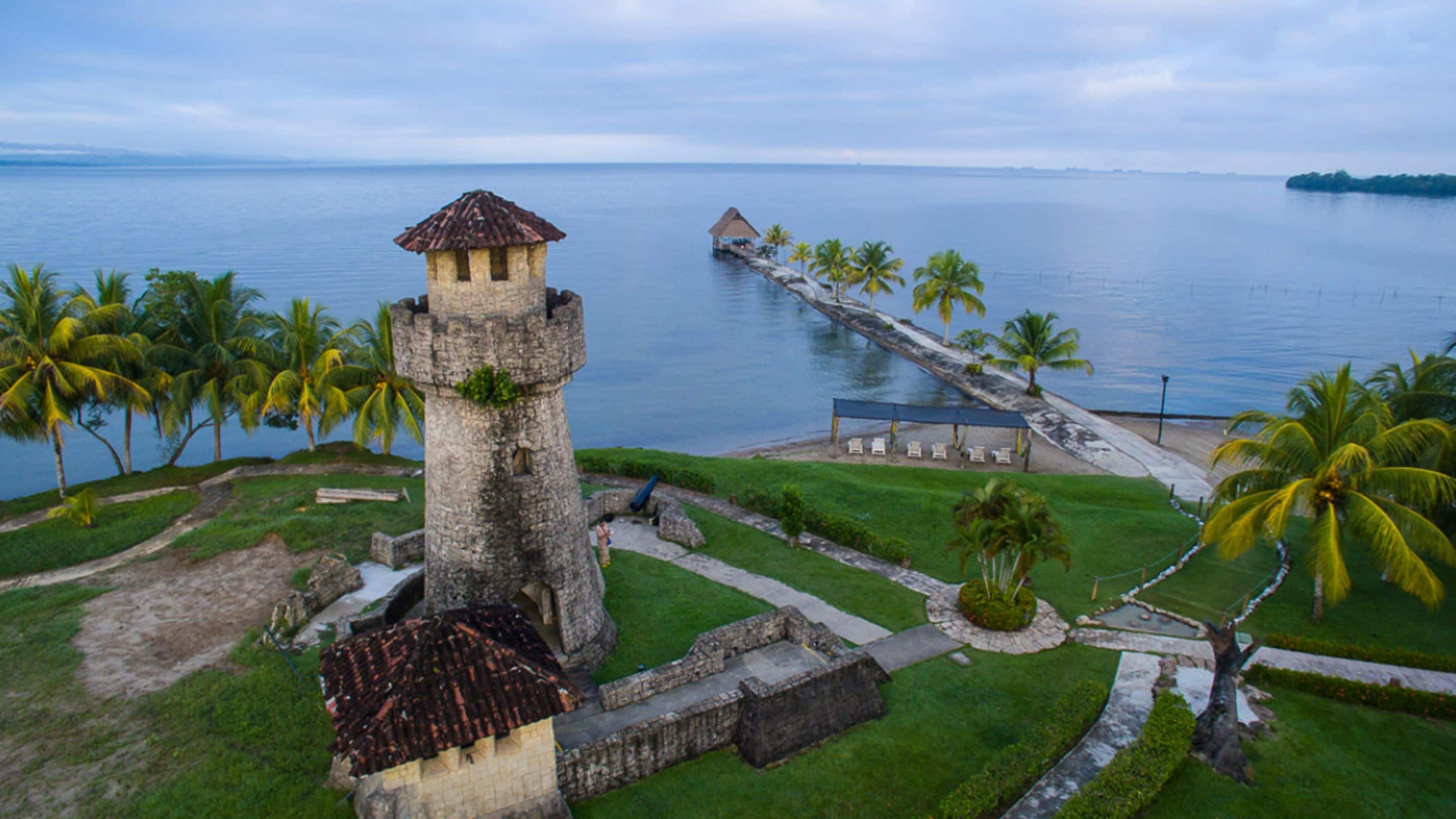 Una fortaleza costera con una torre central, un muelle que se adentra en el agua y una zona de playa con palmeras.