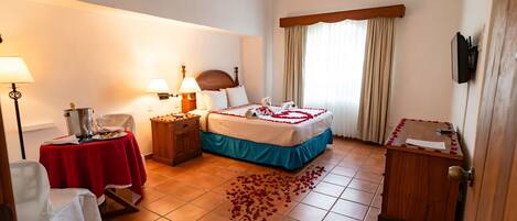 Habitación de hotel con cama, mesita de noche, una mesa con mantel rojo, una silla, un armario de madera y un televisor fijado a la pared.