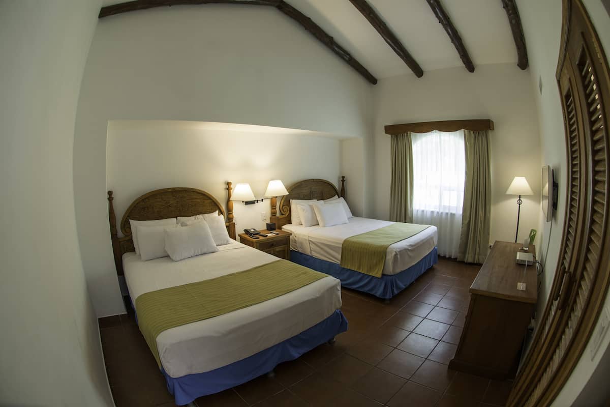 Hotelkamer met twee bedden, houten hoofdeinden, bedlampen, een bureau en een raam met gordijnen.