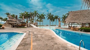 Área de piscina en un resort, con un sendero de piedra que lleva al agua, rodeada de palmeras y una estructura con techo de paja.