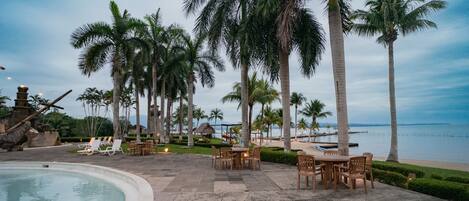 A resort pool area with lounge chairs and tables, palm trees, and a view of the beach and ocean.