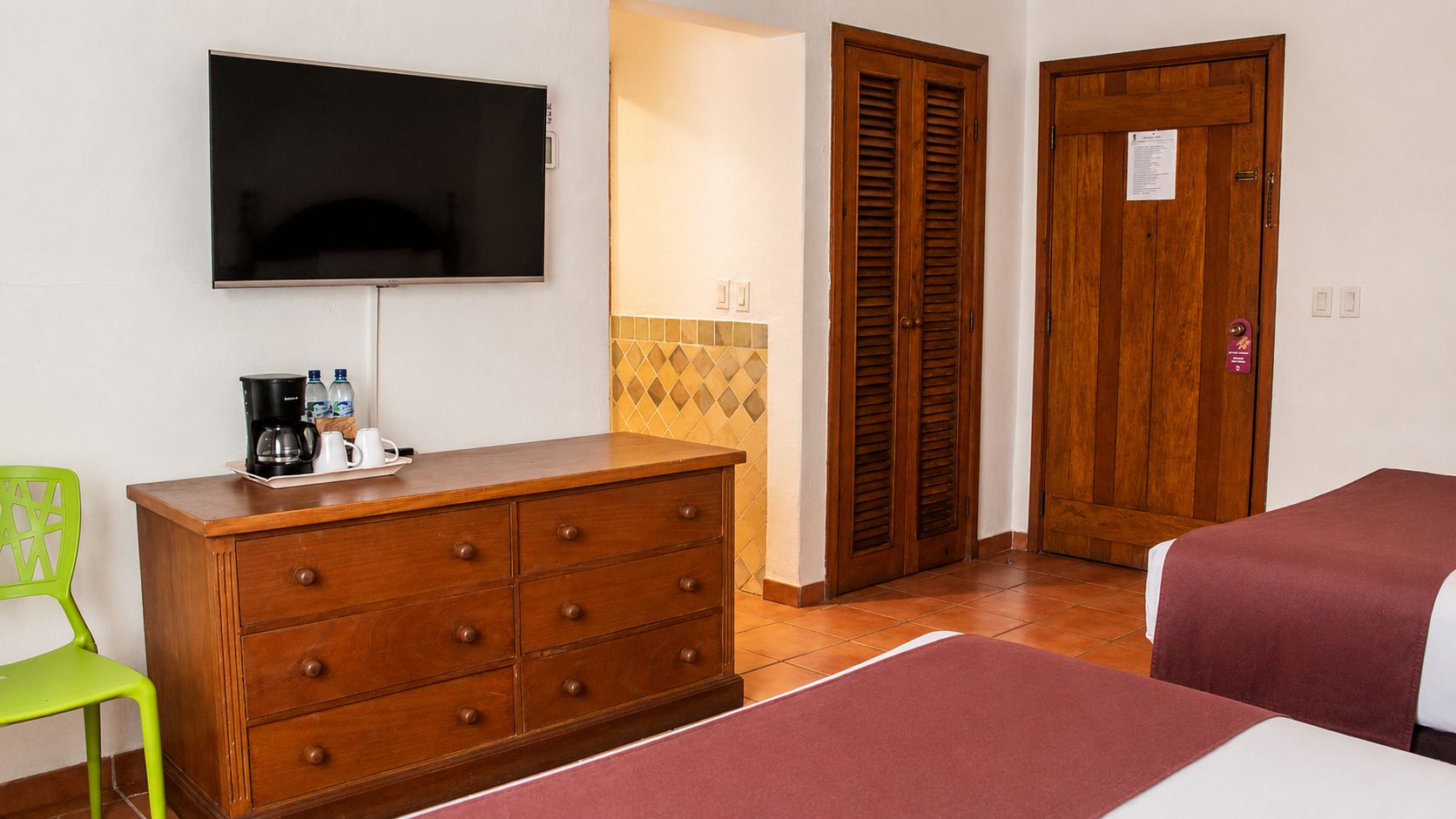 A hotel room with a television, a wooden dresser, a green chair, a bed with a red bedspread, and a wooden door with a sign.