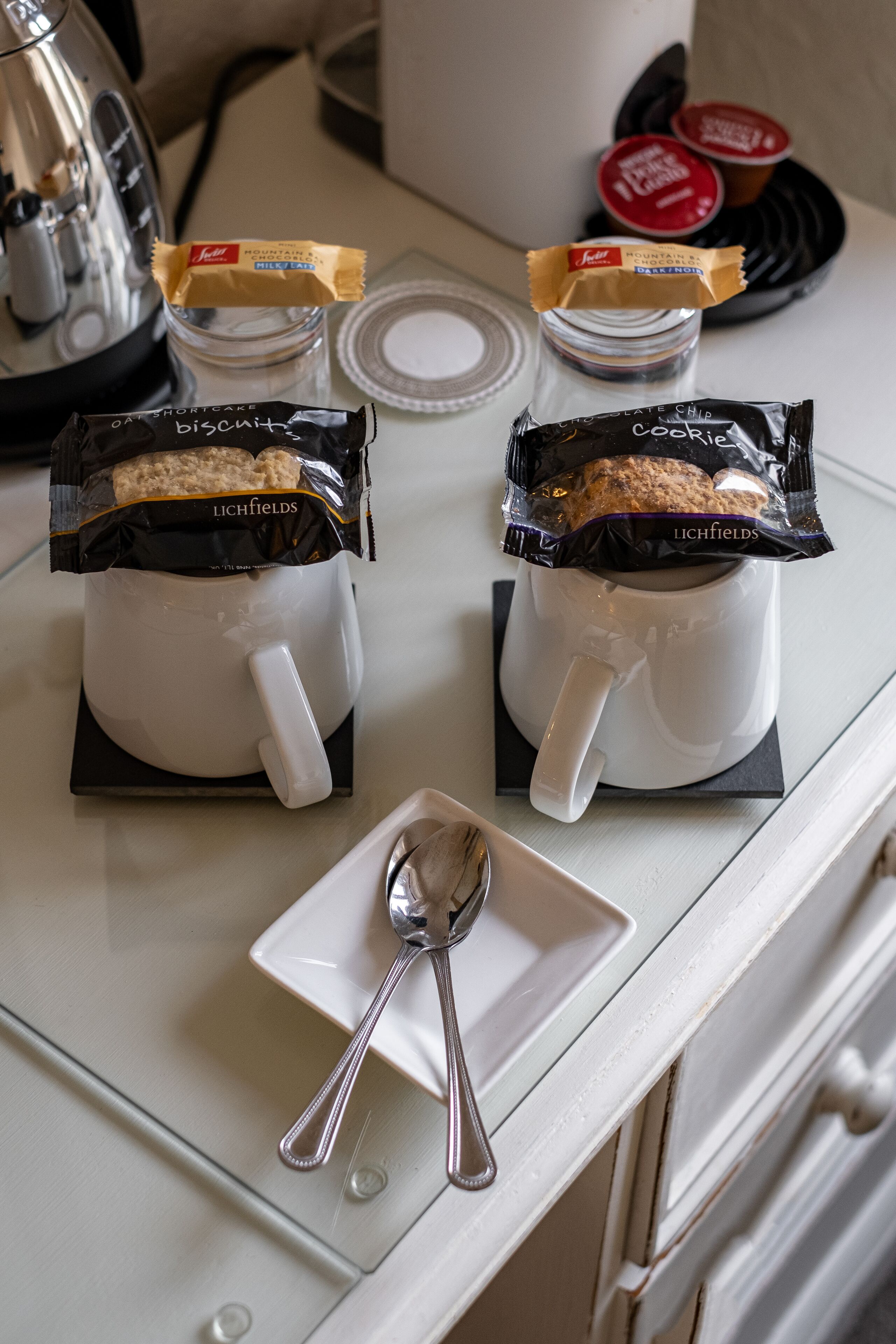 Two white mugs on a table with biscuit packets, a spoon, and a coffee maker in the background.